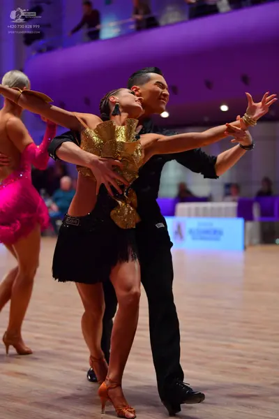 Dancesport competitor: Foreground female dancer: gold sequined and ruffled Latin top with a short black