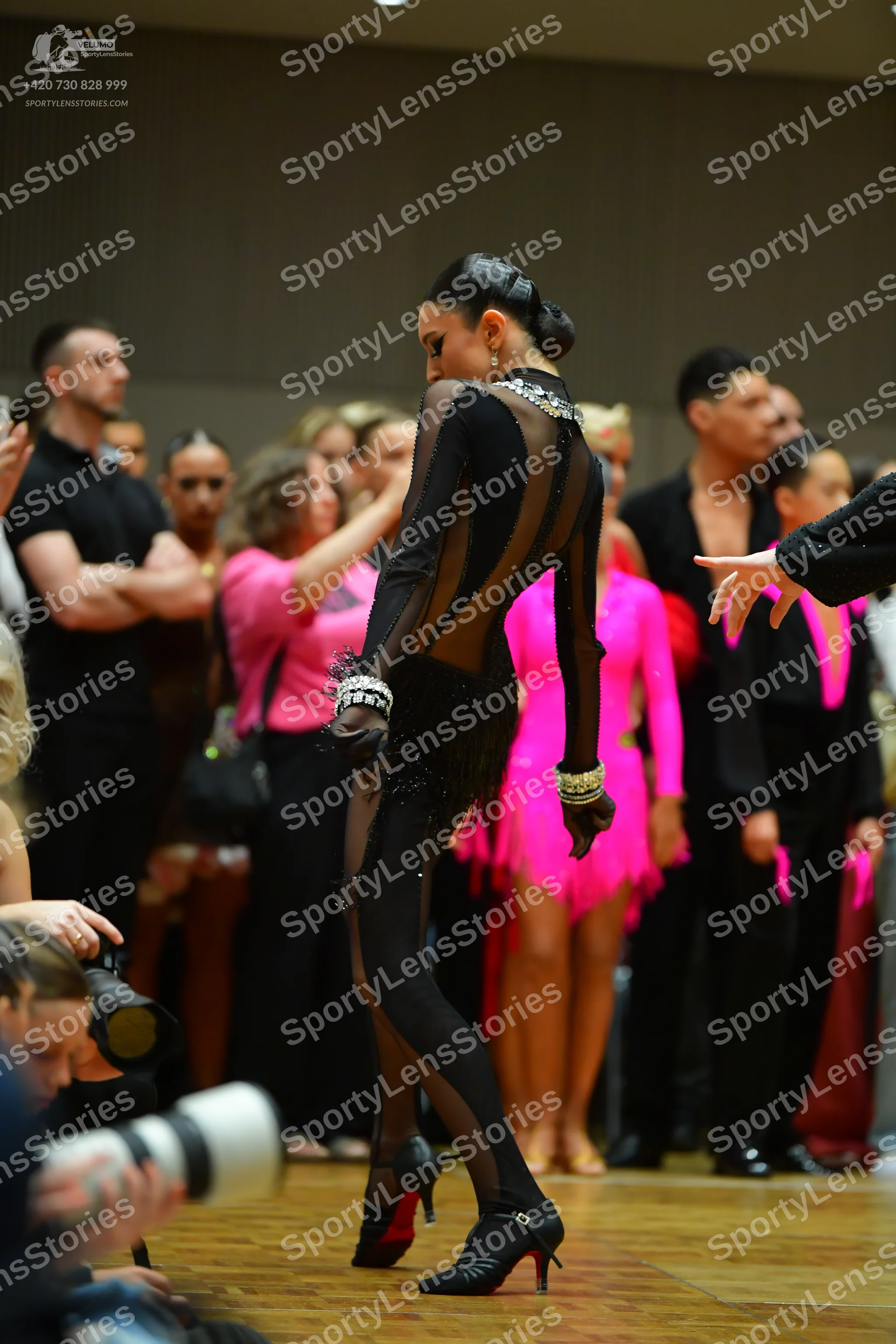 Dancesport competitor: Female dancer in foreground: black Latin dance costume with sheer mesh sleeves a