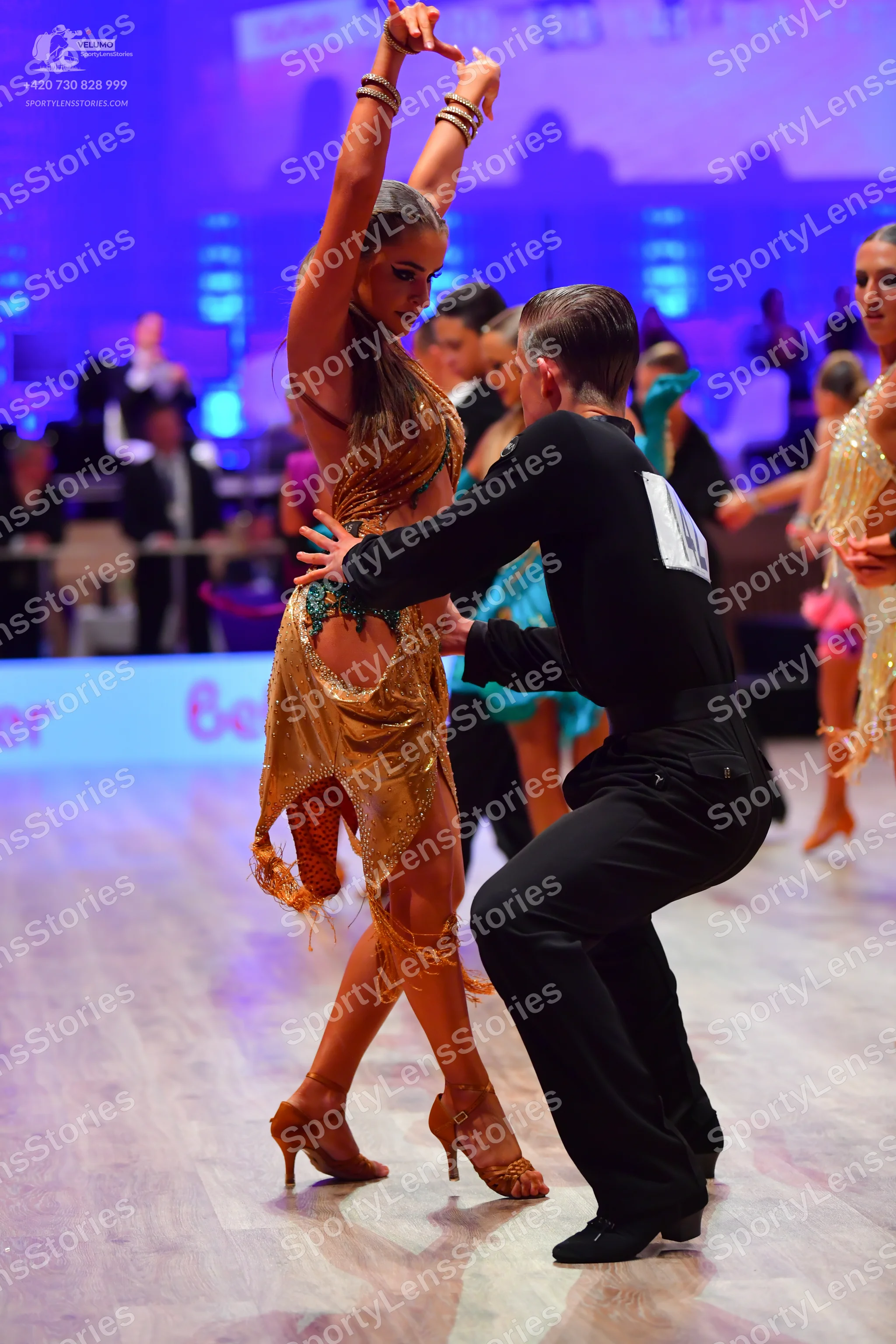 Dancesport competitor: Foreground female dancer: gold/bronze sequined Latin dress with fringe, gold dan