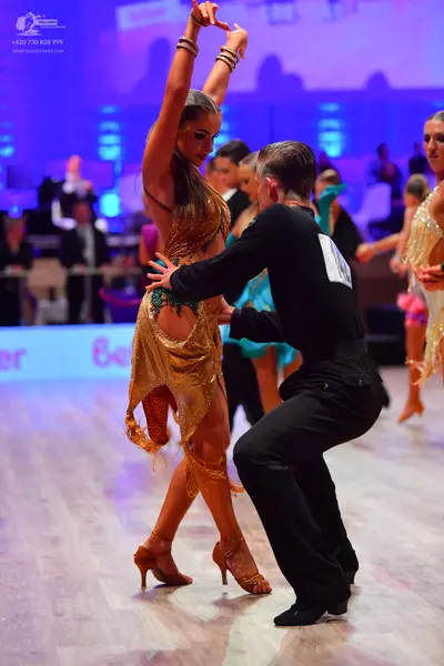 Dancesport competitor: Foreground female dancer: gold/bronze sequined Latin dress with fringe, gold dan