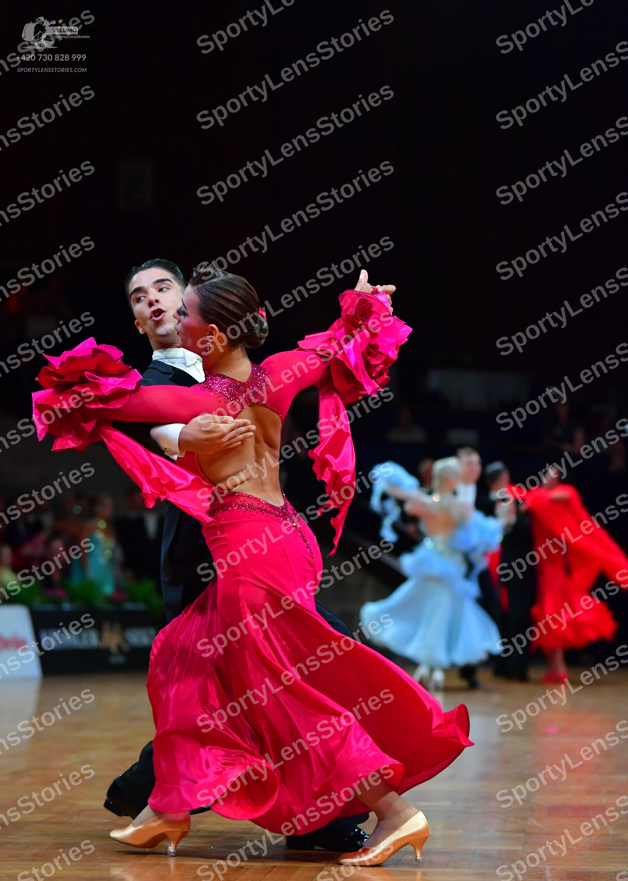 Dancesport competitor: Foreground female dancer: bright pink Latin dress with long ruffled sleeves, a l