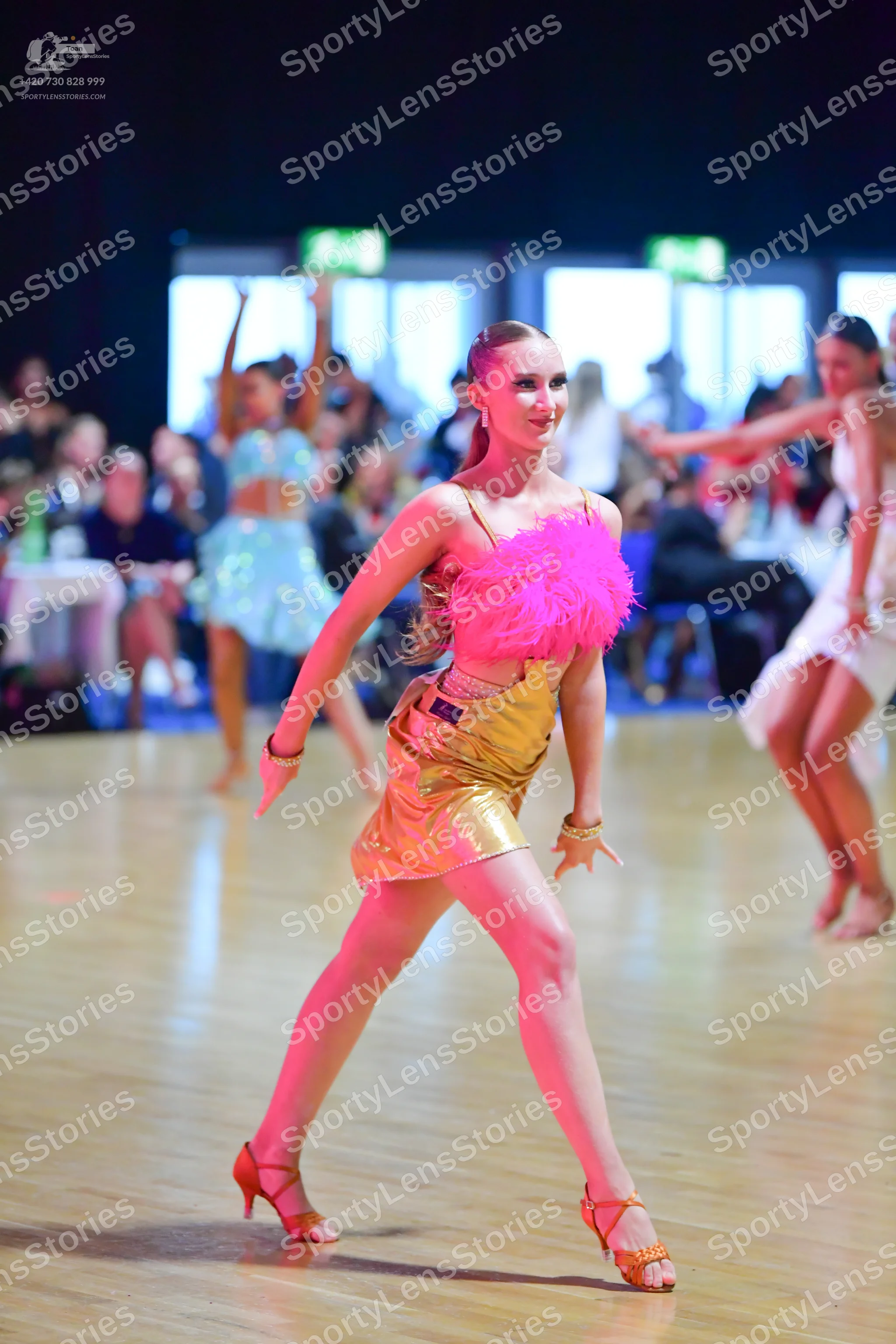 Dancesport competitor: Female dancer in foreground: pink feathery top and a gold metallic skirt, paired