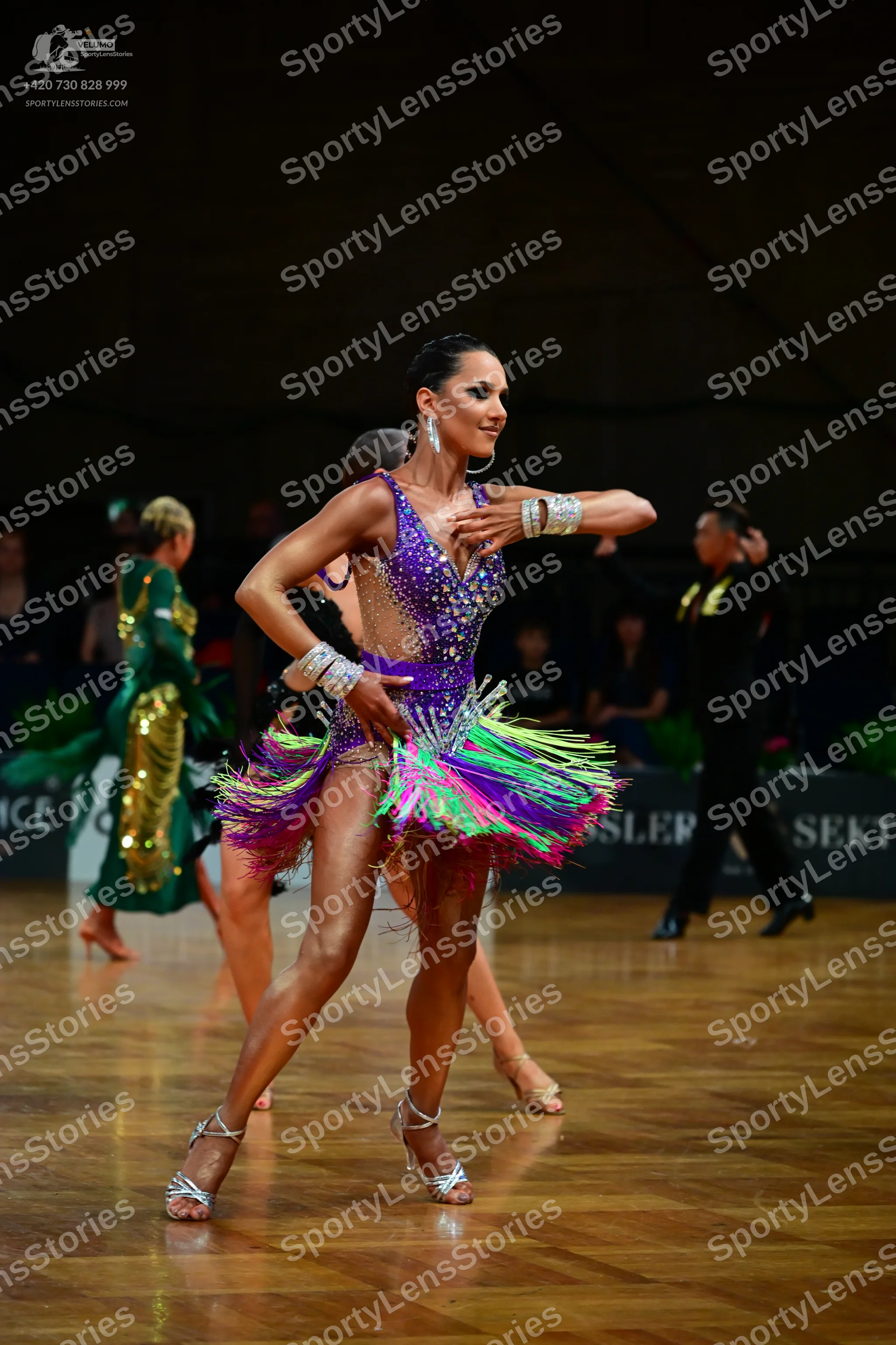 Dancesport competitor: Female dancer in foreground: purple latin dress with colorful fringe (green, pin