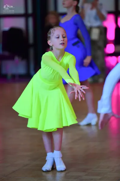 Dancesport competitor: Female dancer in foreground: neon yellow long-sleeved Latin dress with a flared 